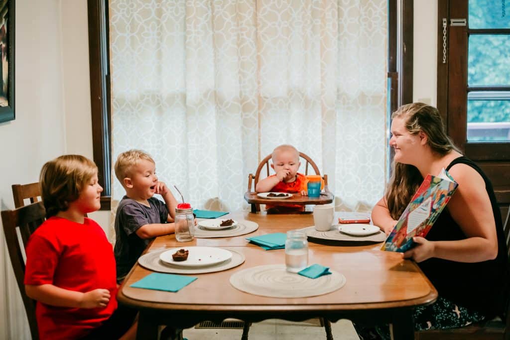 Mom and 3 kids sitting around the table, eating breakfast, and reading a book.