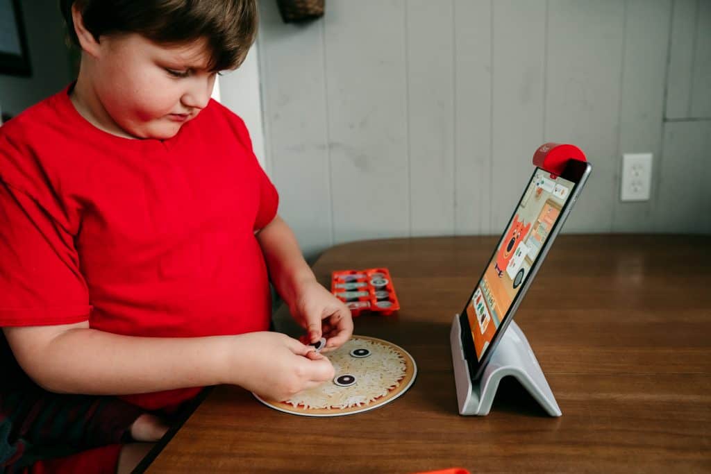 Boy playing Osmo Pizza Co game on an iPad with hands-on game pieces.
